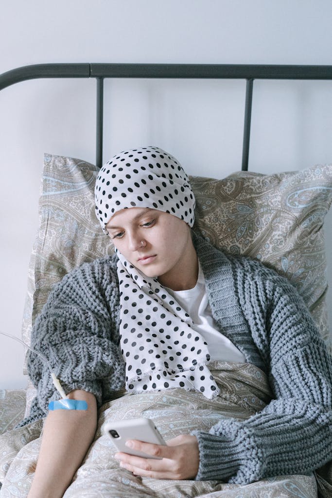 Woman with headscarf using a smartphone in bed, receiving cancer treatment.